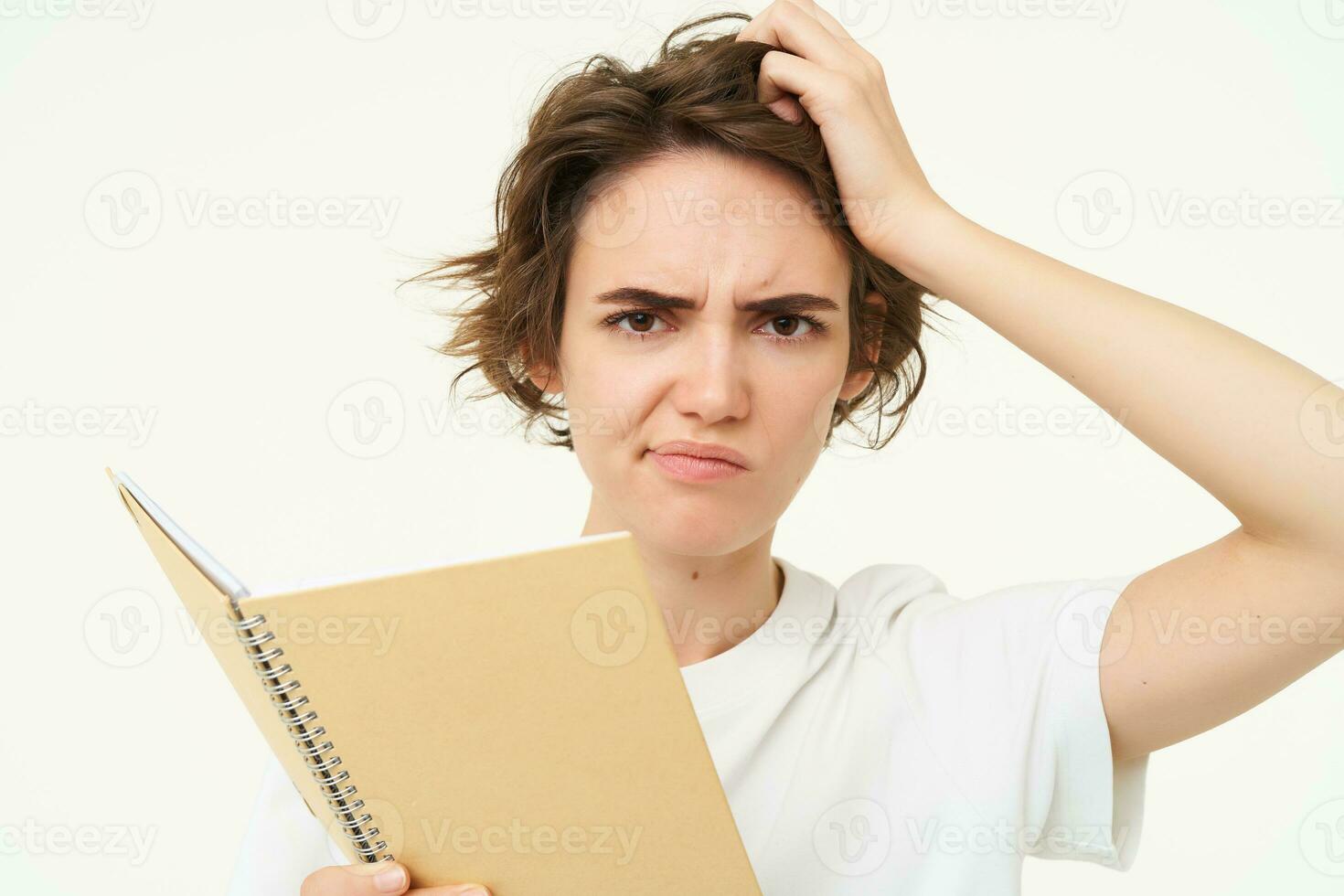 Portrait of confused girl scratching her head, holding planner, student doing homework with puzzled face, thinking with frowning expression, standing over white background photo