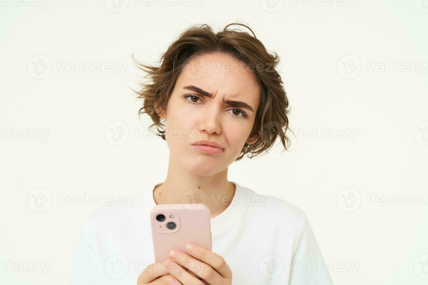 Image of complicated, confused young woman, frowning, holding smartphone, looking puzzled and doubtful, standing over white background photo