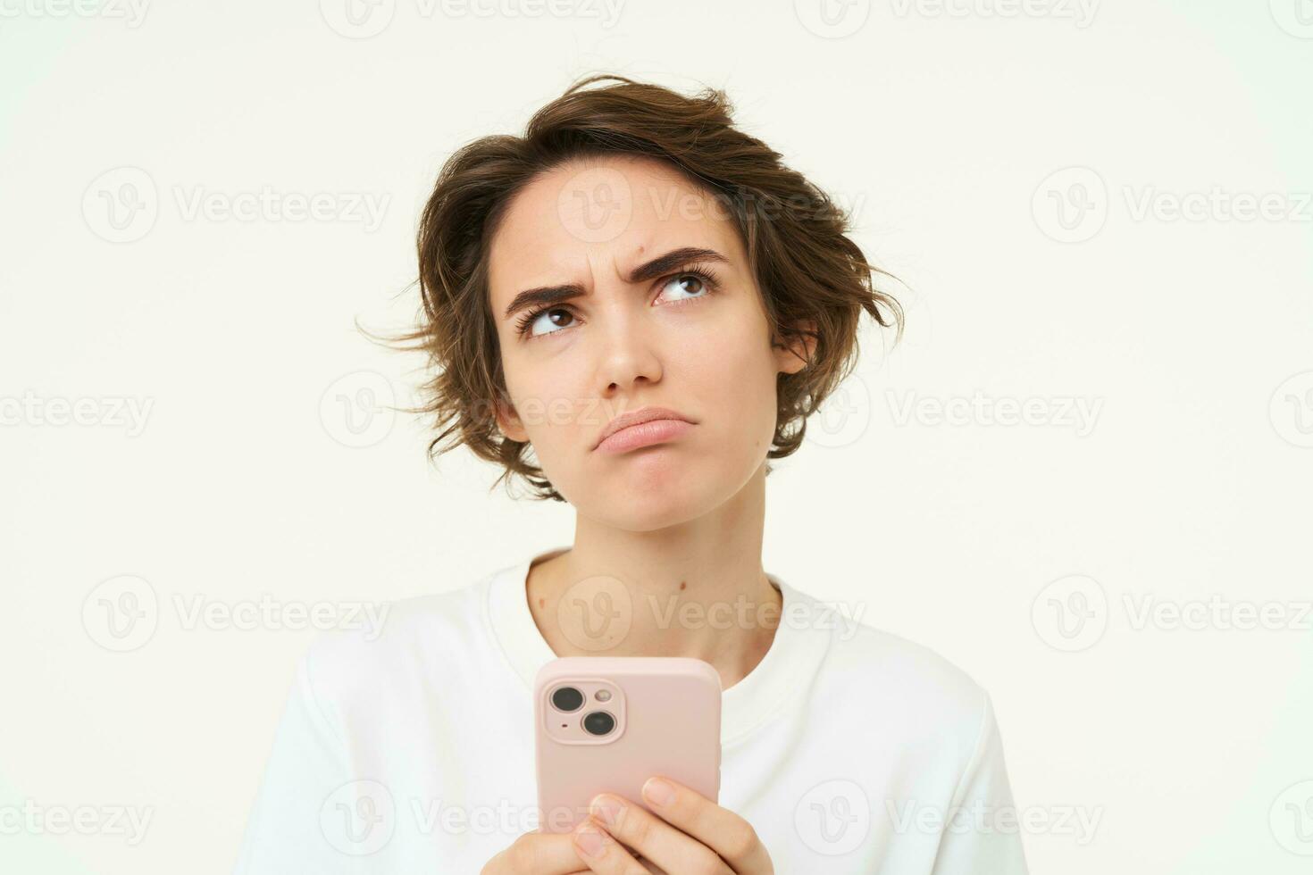 Portrait of puzzled brunette woman, standing with smartphone, thinking and frowning, looking reluctant, standing over white background photo