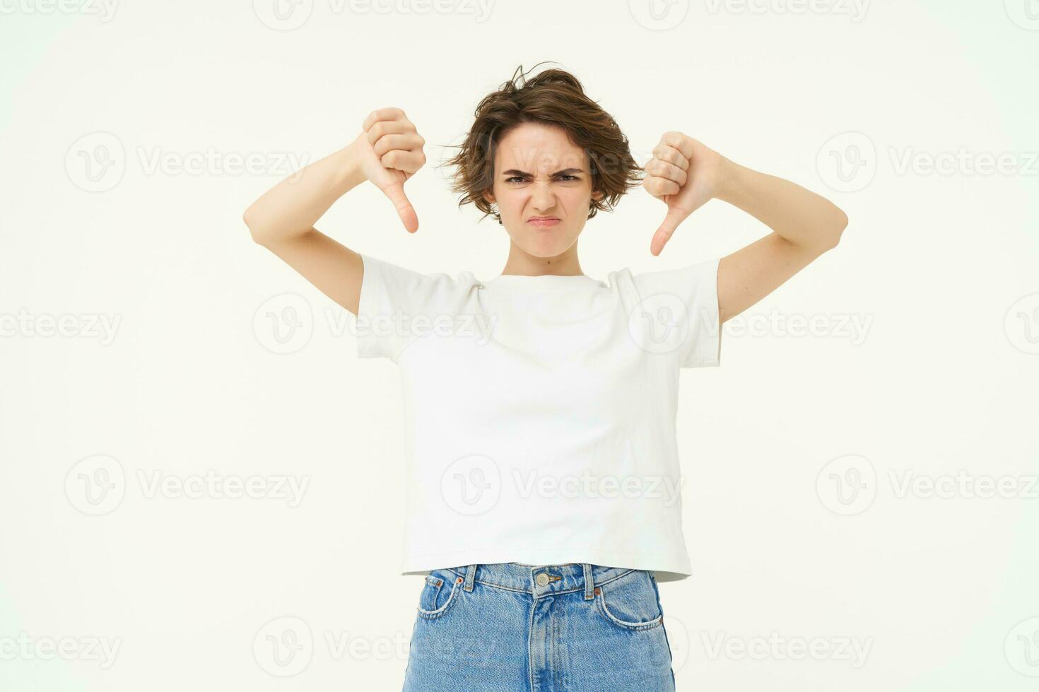 Portrait of disappointed young woman, female client shows dislike gesture, thumbs down, stands over white background photo