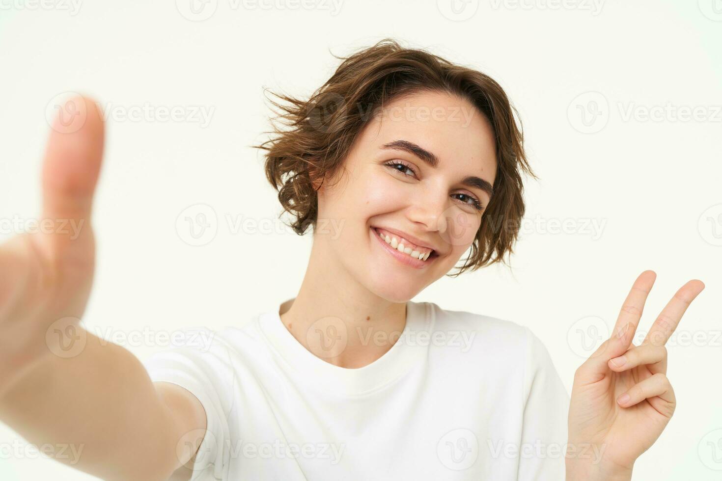 cerca arriba de sonriente morena mujer hace autofoto, tomando foto para su social medios de comunicación, sostiene móvil teléfono con extendido mano, en pie terminado blanco antecedentes