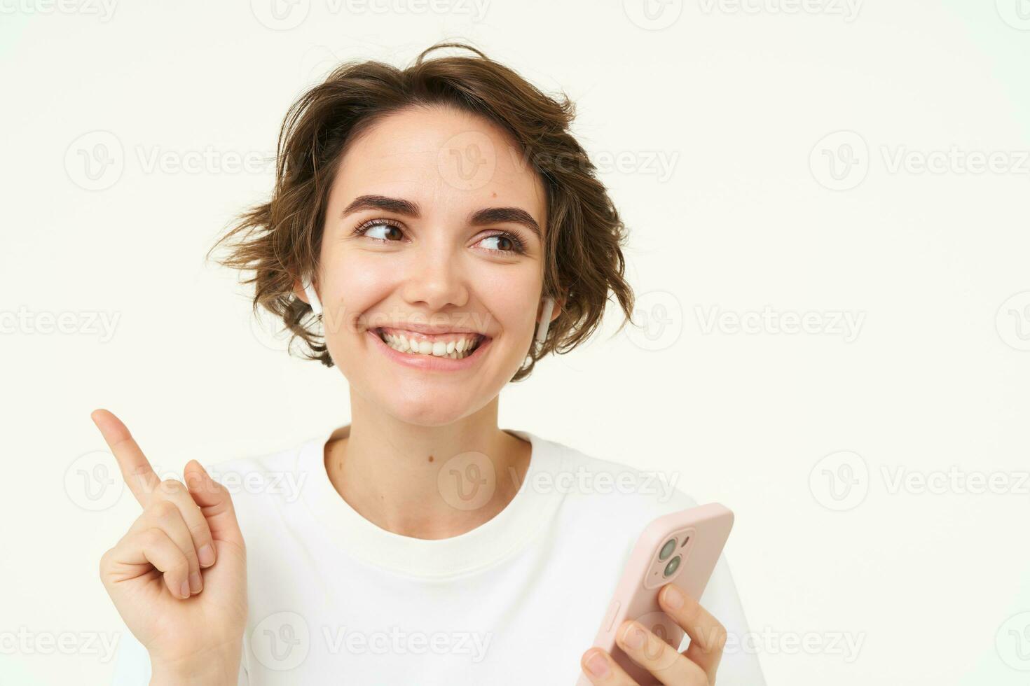 Portrait of brunette woman listening music in headphones, pointing left at banner, holding mobile phone, using streaming app, isolated over white background photo