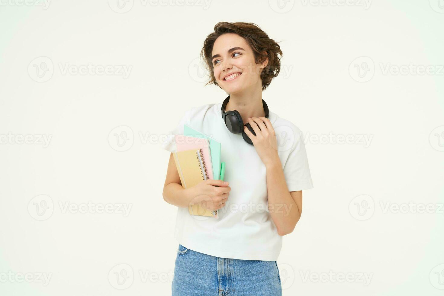 Image of brunette woman in headphones, standing with documents. Student with notes and workbooks posing against white background photo