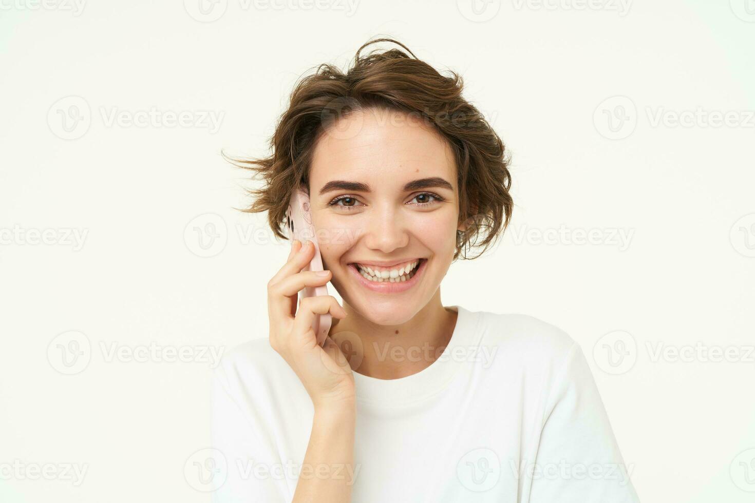 Image of cute brunette woman answer phone call, talking on smartphone, holding mobile near eat and smiling , standing over white background photo