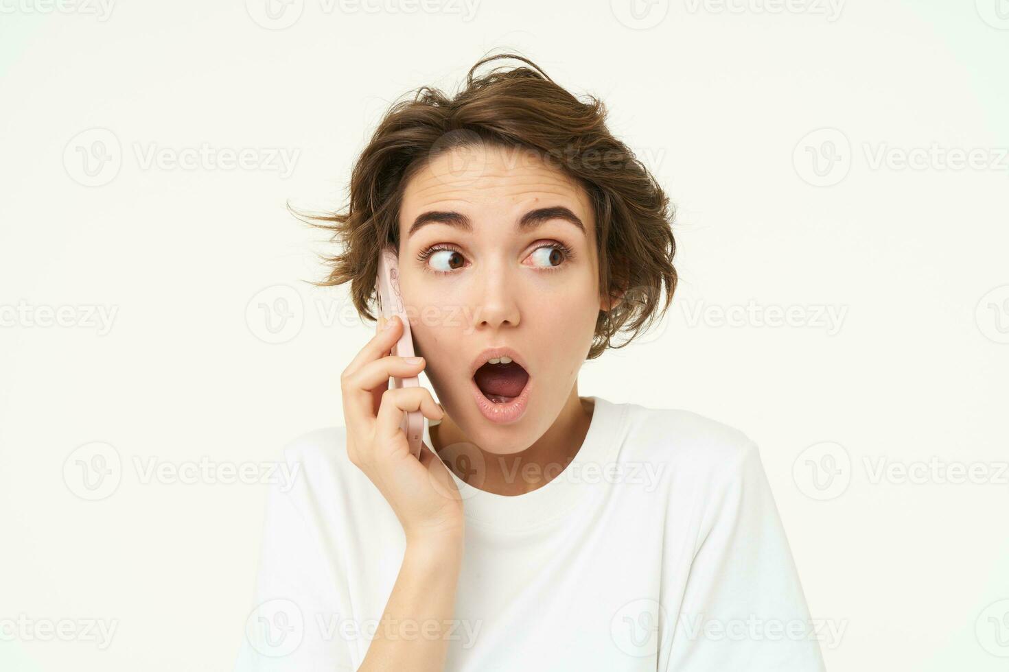 Portrait of amazed brunette girl looks surprised while talks on mobile phone, answers a call, amazed by big news, gasping shocked, standing over white background photo