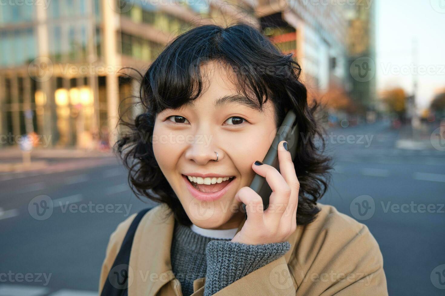 Portrait of happy korean girl, talks on mobile phone, looks surprised and happy, receives positive great news over telephone conversation, stands on street of city photo