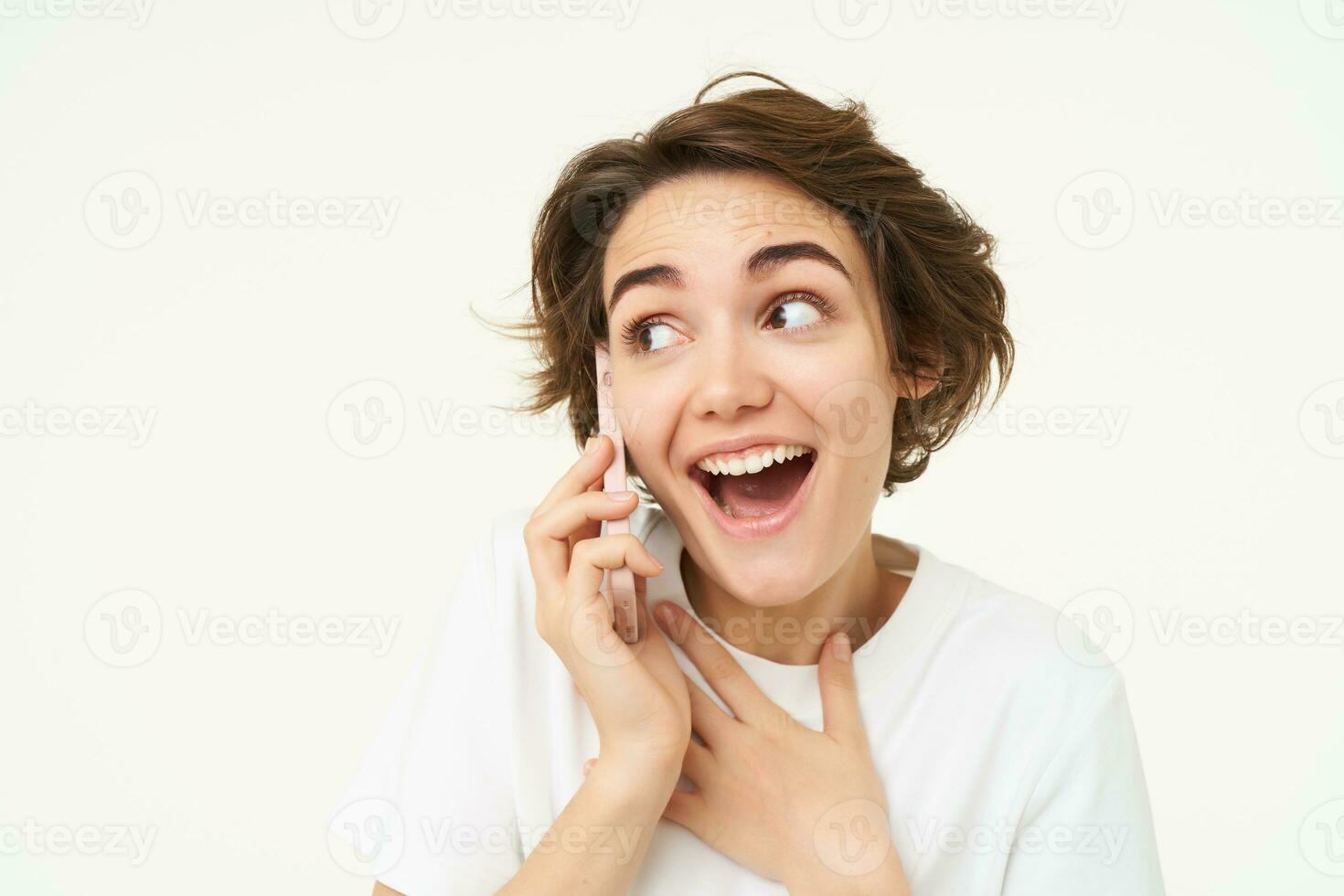 Portrait of amazed brunette girl looks surprised while talks on mobile phone, answers a call, amazed by big news, gasping shocked, standing over white background photo