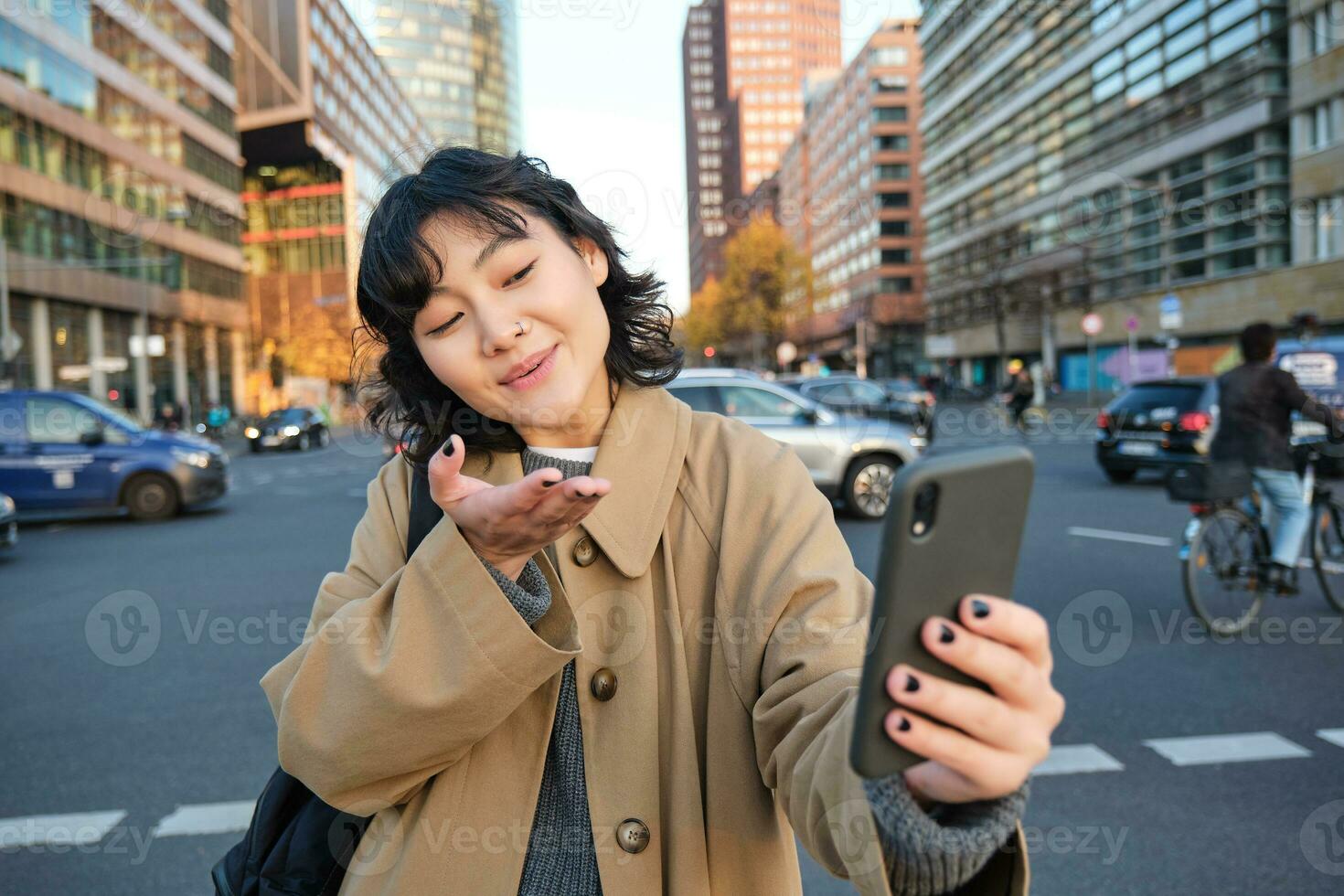 Cute korean girl, tourist takes selfie, video chats with friend and sends kisses to camera, records vlog on streets of city with smartphone photo