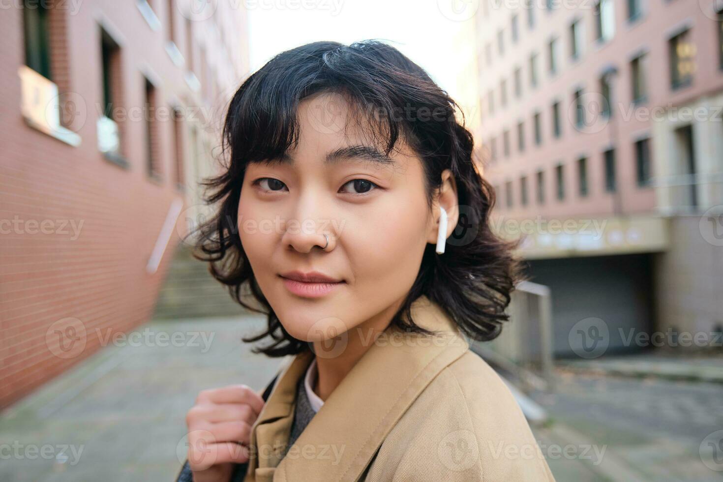 Stylish modern asian girl, student in wireless headphones, walks around city with backpack, smiling with happy face photo