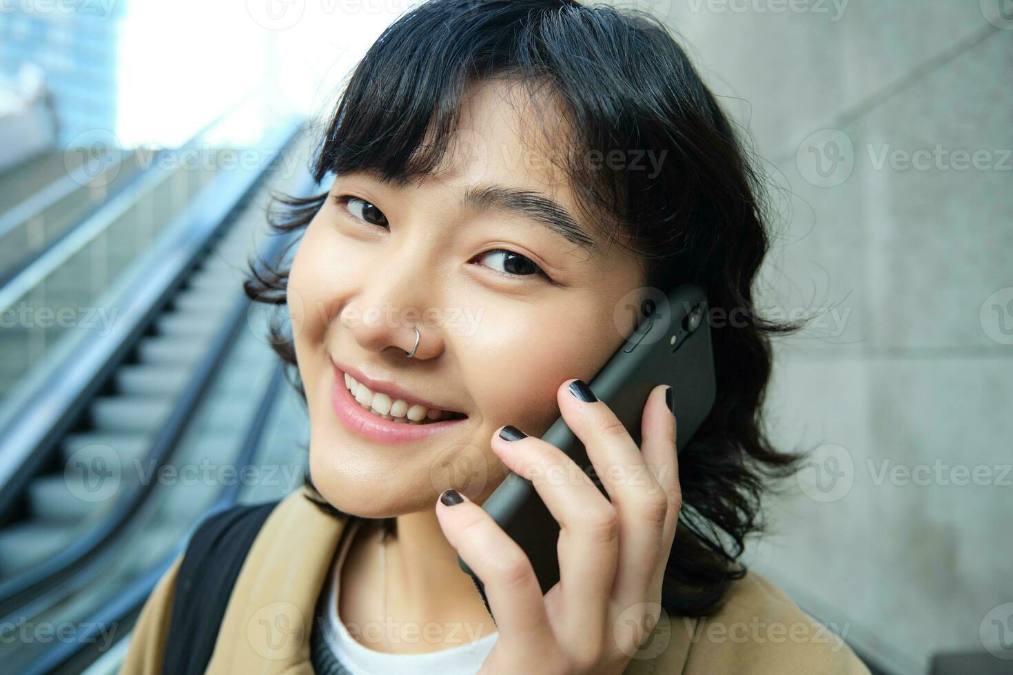 Close up portrait of smiling brunette girl, korean woman goes down escalator, makes phone call, talks to someone on telephone photo