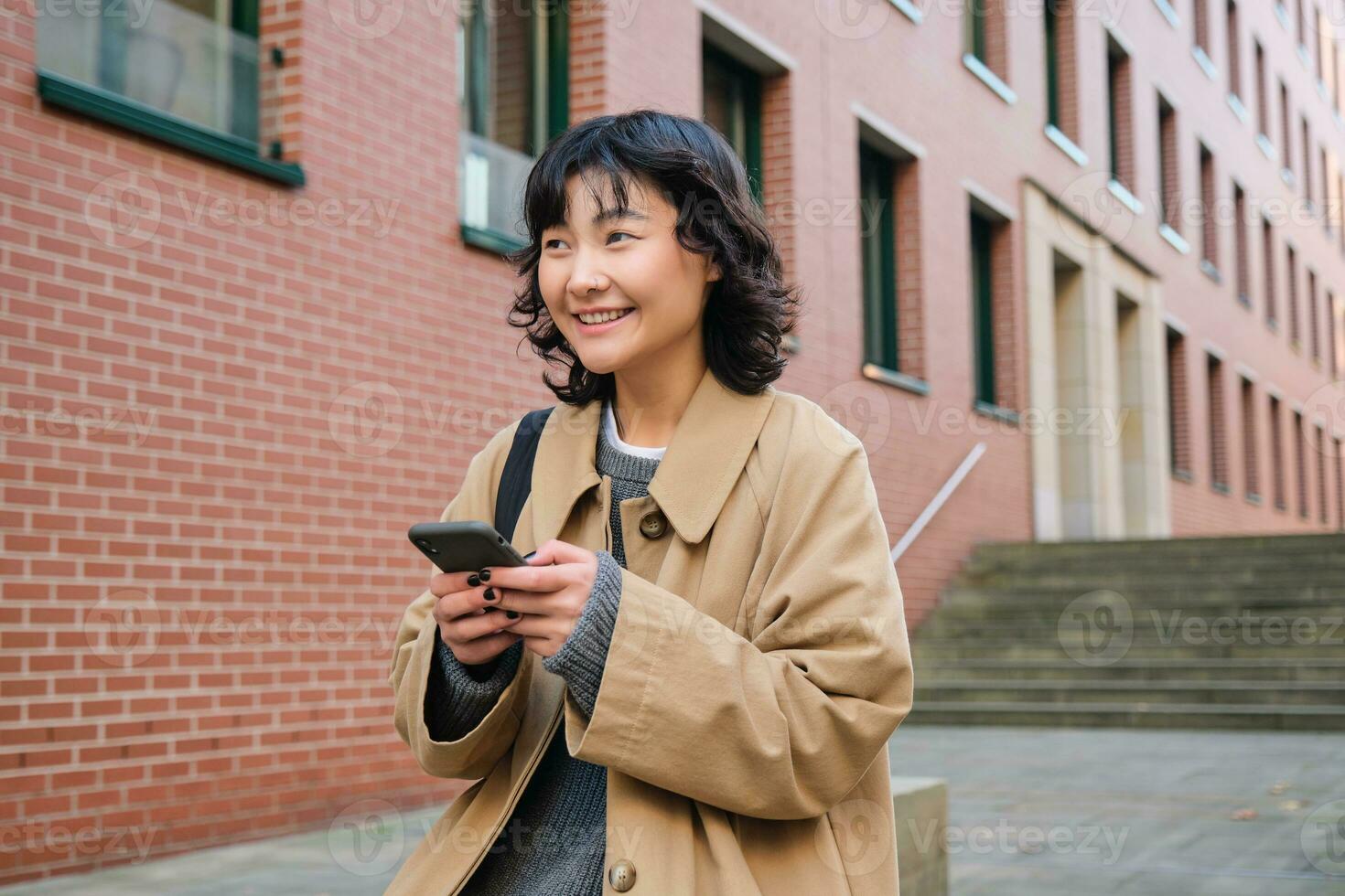 Stylish korean hipster girl, walks in city centre, sends text message, uses location map application on smartphone while walking down the street, wears trench coat photo