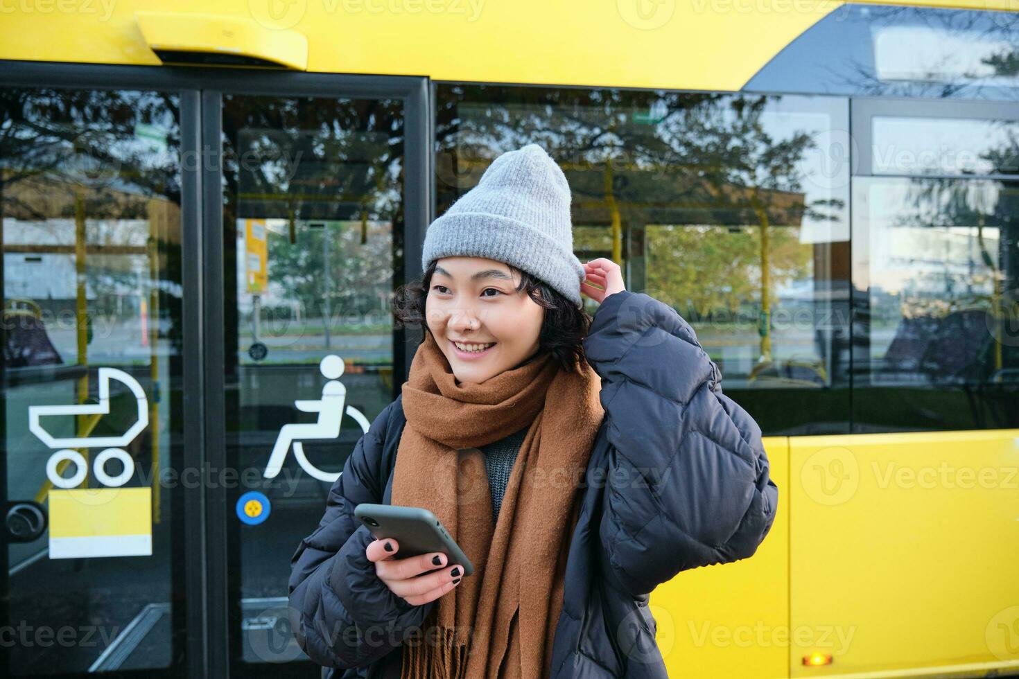 retrato de niña en pie cerca autobús en un detener, esperando para su público transporte, cheques calendario en teléfono inteligente solicitud, sostiene móvil teléfono, usa calentar ropa foto