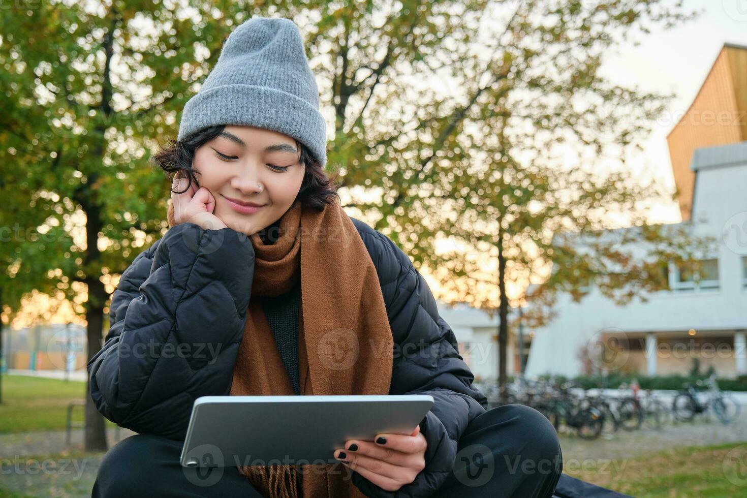 Smiling asian girl, student sits on bench in park alone, reading, using social media app on digital tablet, watches videos outdoors, relaxes on fresh air photo