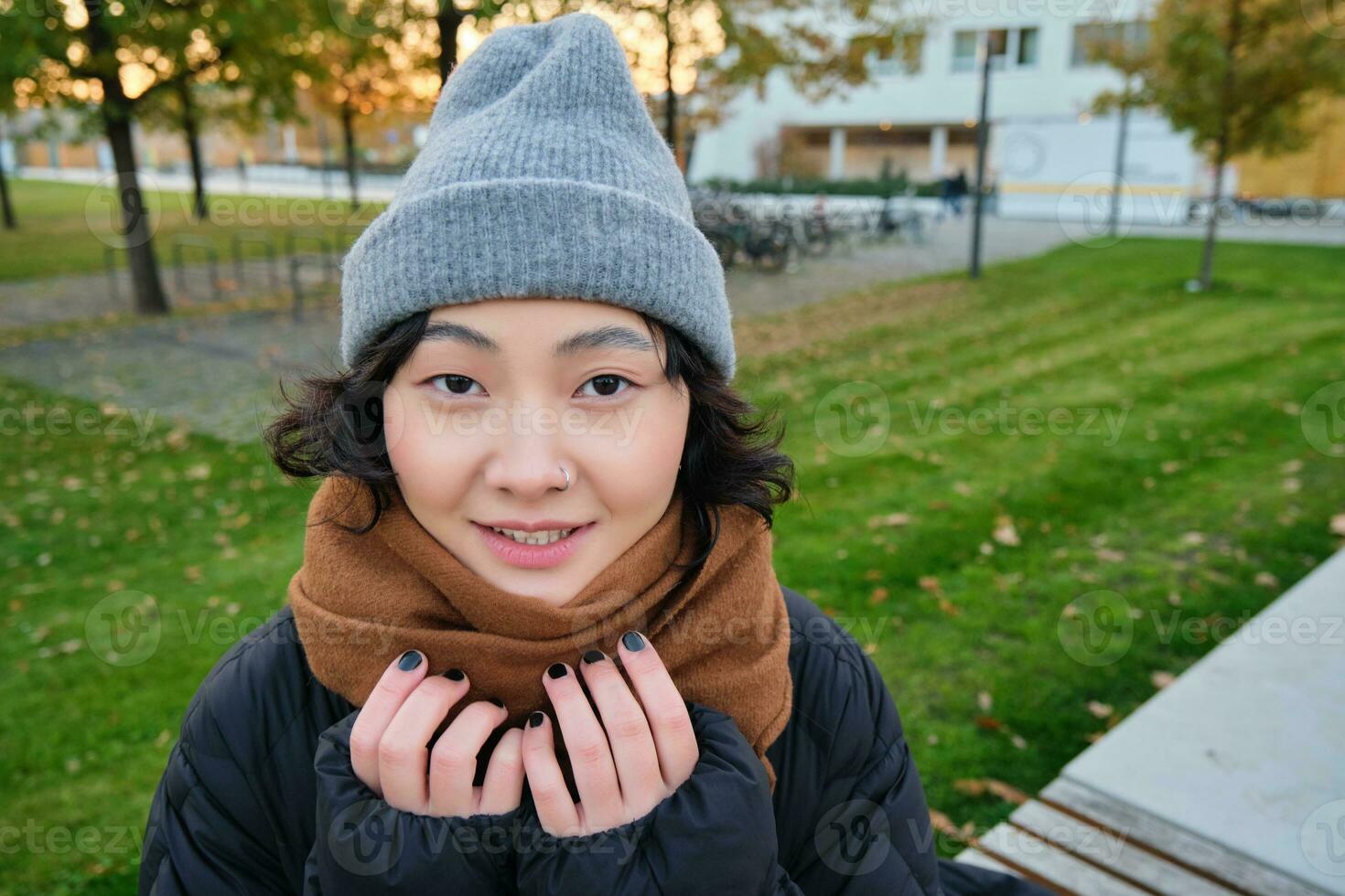 Portrait of cute asian girl in hat and scarf, walks around town in chilly spring weather, smiles and looks happy, sits in park near green grass photo