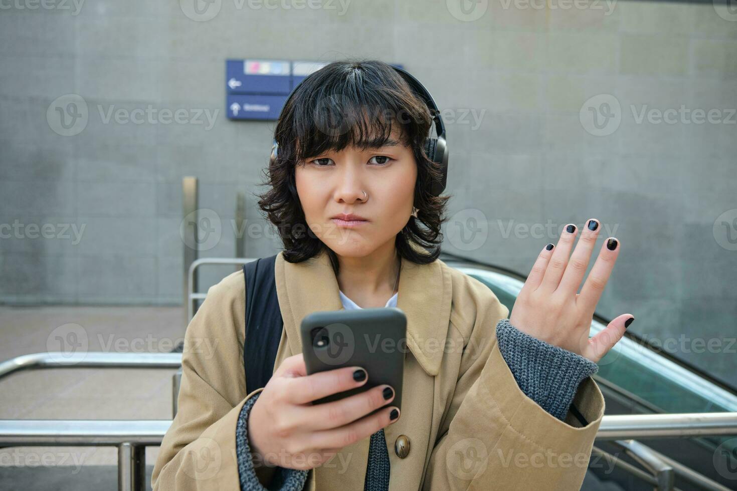 Portrait of asian girl in headphones, looks complicated at smartphone screen, puzzled by text message or notification, stands on street and shrugs photo