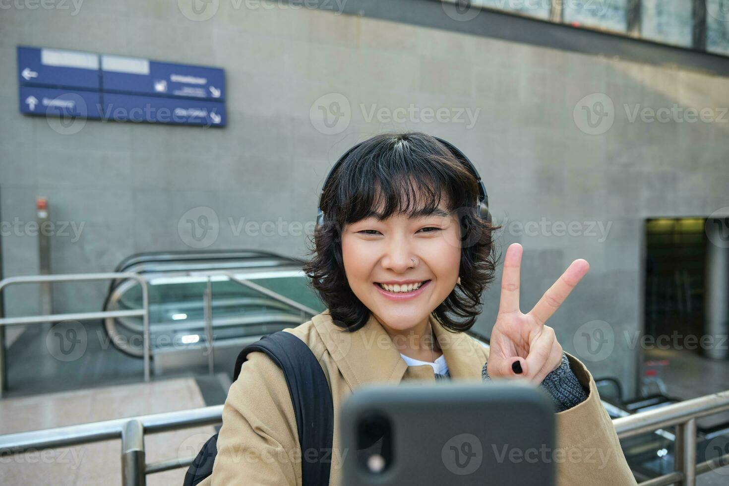 linda y elegante coreano chica, usa auriculares, toma selfie en teléfono inteligente, turista registros vídeo o hace un foto, soportes en calle foto