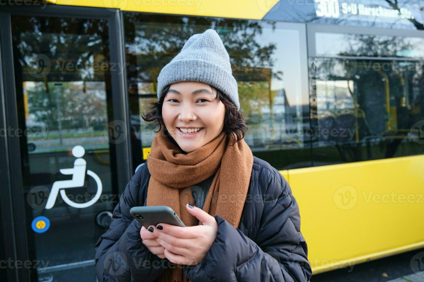 imagen de niña estudiante esperando para público transporte, cheques calendario en teléfono inteligente aplicación, soportes cerca ciudad autobús foto