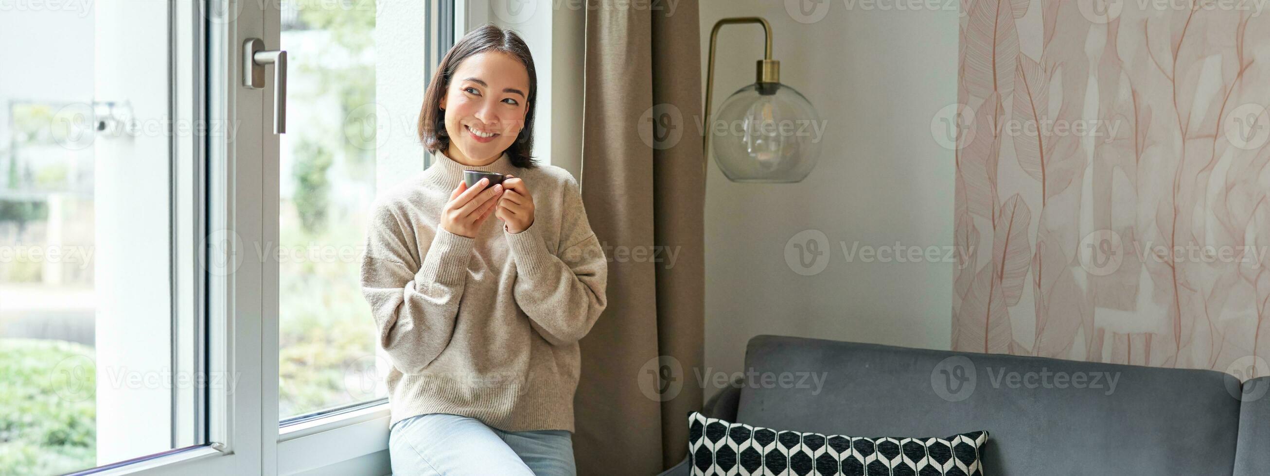 Beautiful young asian woman sitting near window and drinking her coffee, holding espresso cup and looking outside with relaxed, smiling face expression photo