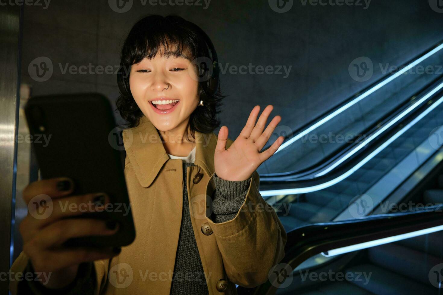Cute girl video chatting on smartphone, standing near escalator and waving hand at mobile phone camera, saying hi while talking to friend via telephone app, wearing headphones photo