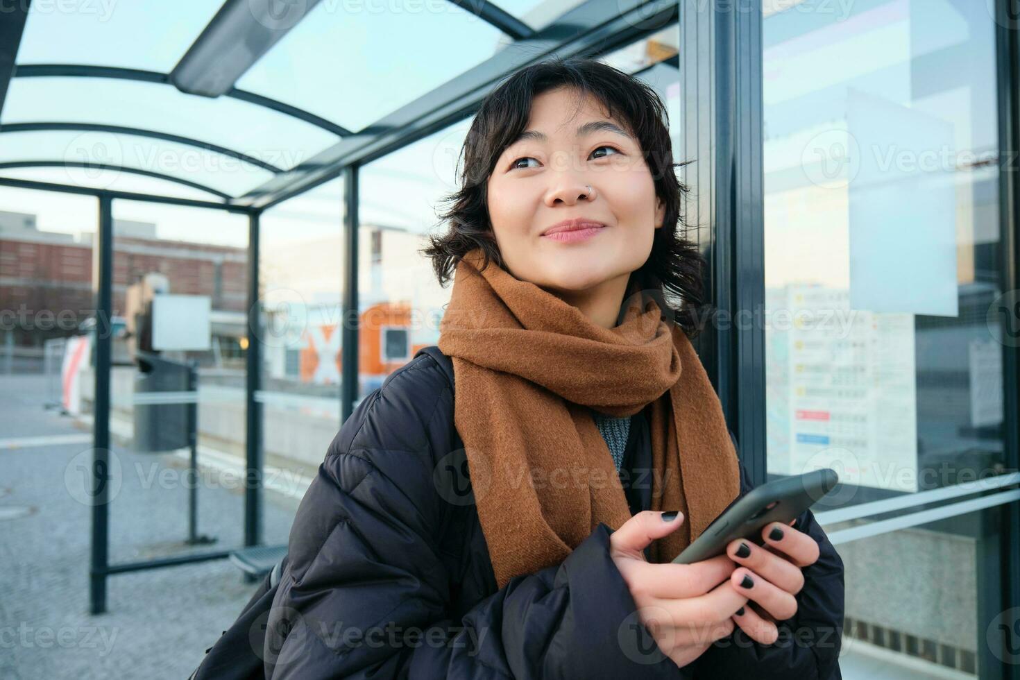 Beautiful Japanese girl, student standing on bus stop in winter jacket, holding smartphone, using application to track her public transport, ordering a ride photo