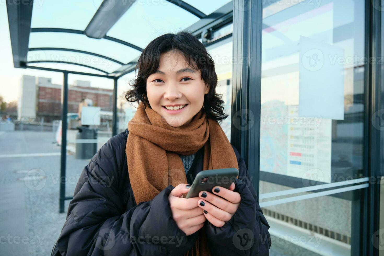 Cute young girl, student waiting on a bus stop, looking for her public transport on app tracker, holding smartphone, standing on street near road photo