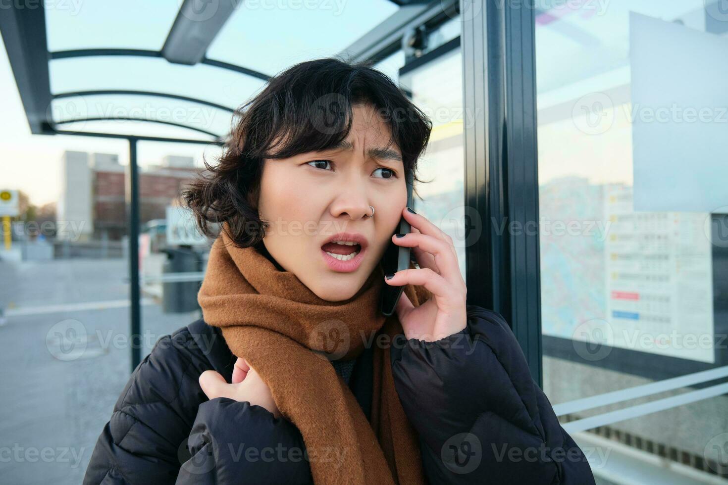 Young woman looks frustrated and disappointed, talks on mobile phone, stands on bus stop, receives bad news photo