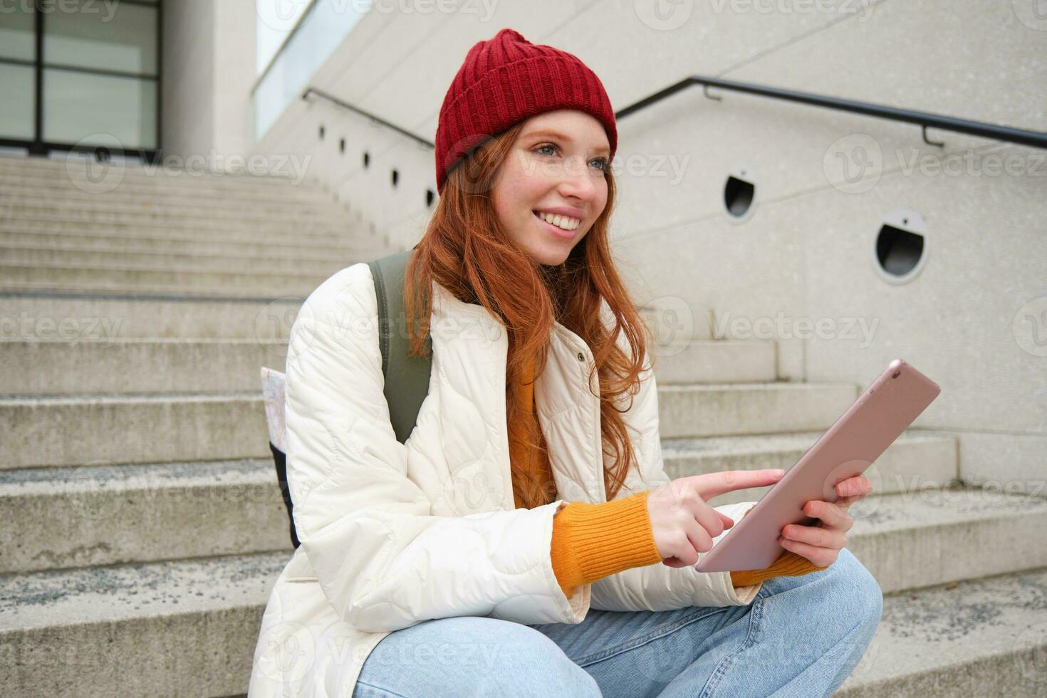 Happy stylish redhead girl, student in red hat, holds digital tablet, uses social media app, searches something online, connects to wifi photo