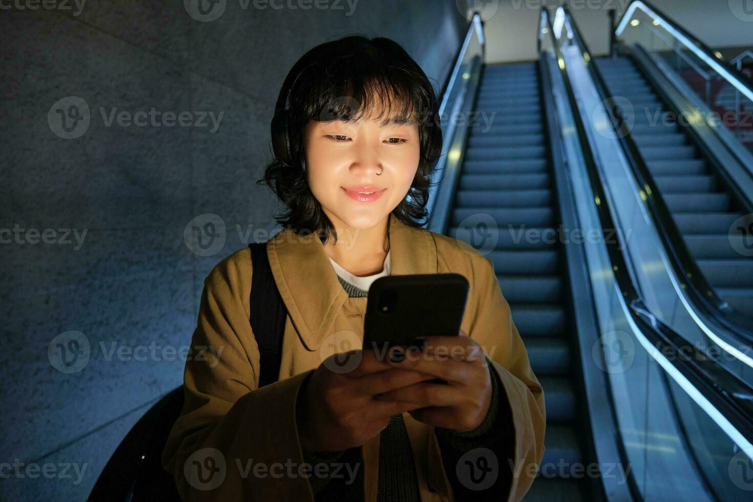 Happy smiling young woman, standing on escalator, going down, holding smartphone in both hands, chatting on mobile phone app photo