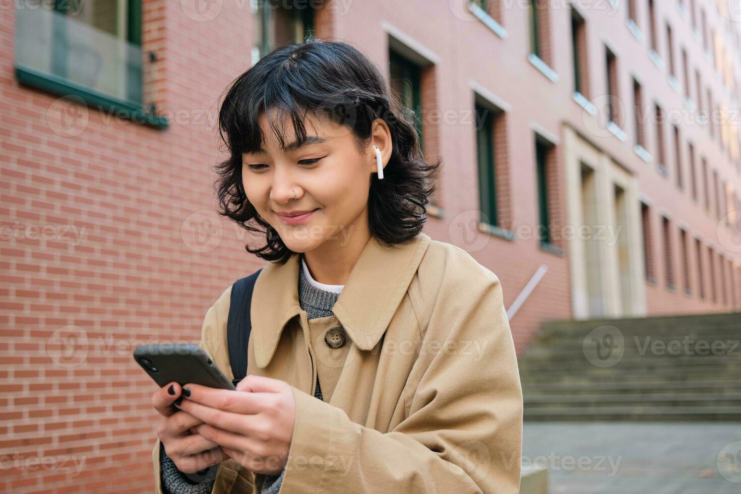 Happy korean girl walks on street, listens music in wireless earphones and holds smartphone, picks song in playlist while standing outdoor near building, reading message photo