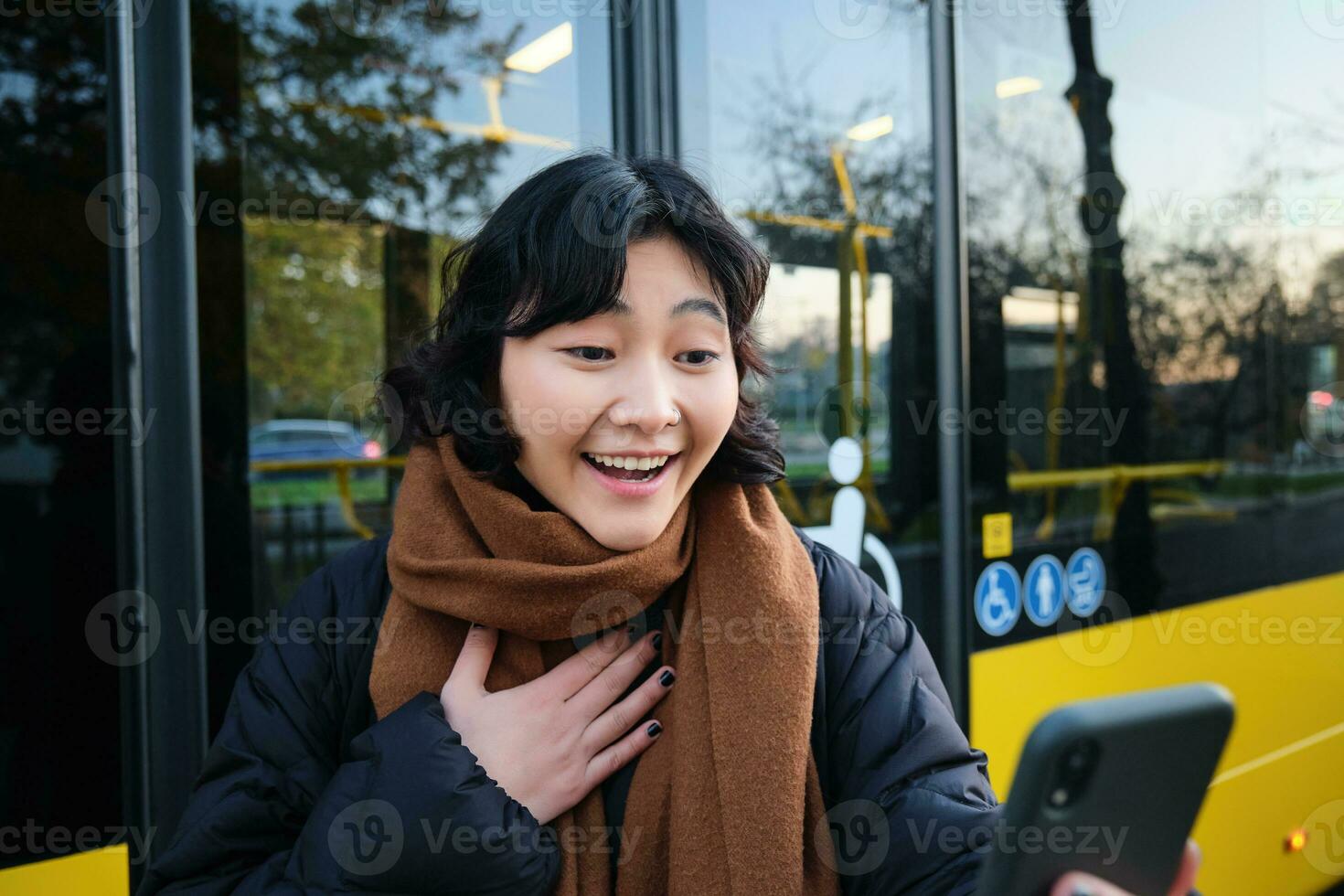 Portrait of cheerful asian girl talks on mobile phone, video chats, looks amazed at smartphone camera, stands on bus stop photo