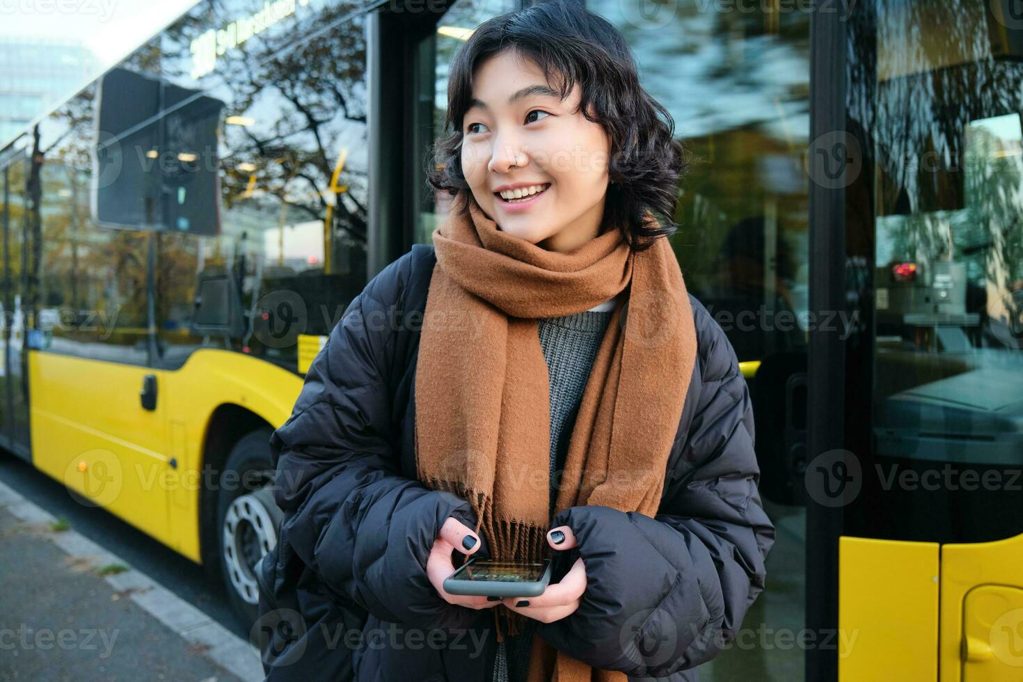 Young beautiful woman standing on bus stop, texting message on smartphone, holding mobile phone, checking her schedule, buying ticket online, wearing winter clothes photo