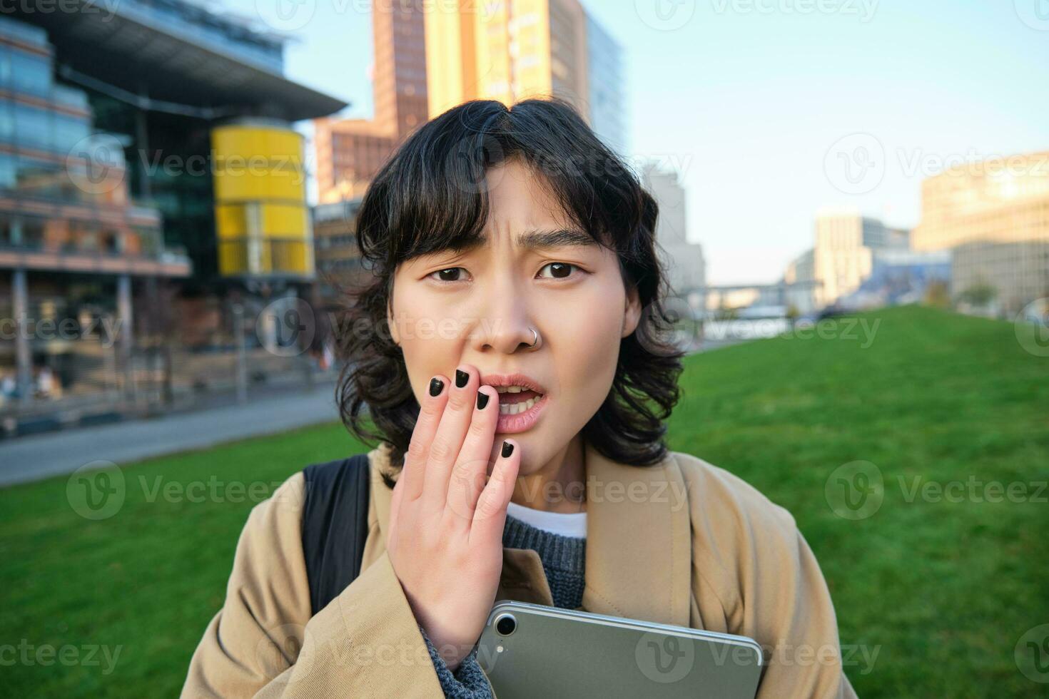 Close up portrait of girl with tablet, looks worried, concerned face expression, feels sad for someone, hears bad news, stands on street near university campus photo