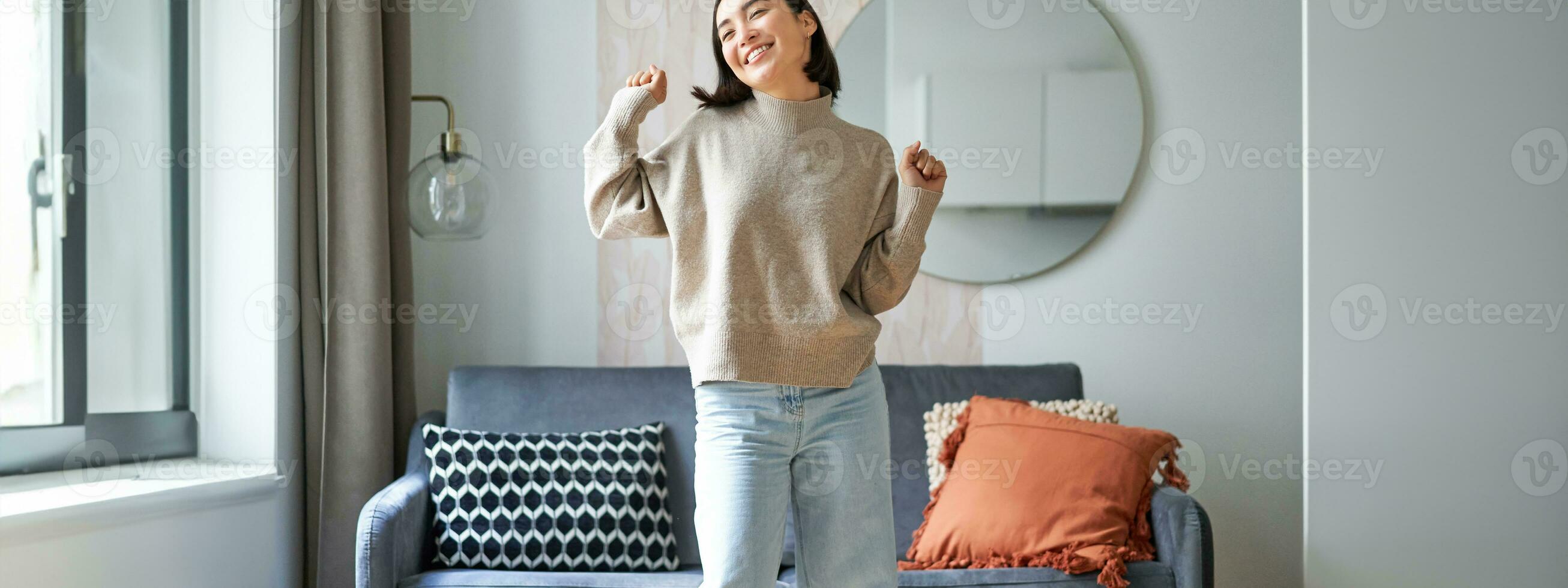 Enthusiastic asian woman dancing, feeling free and upbeat at her home, enjoying happy day, staying indoors, smiling and laughing photo