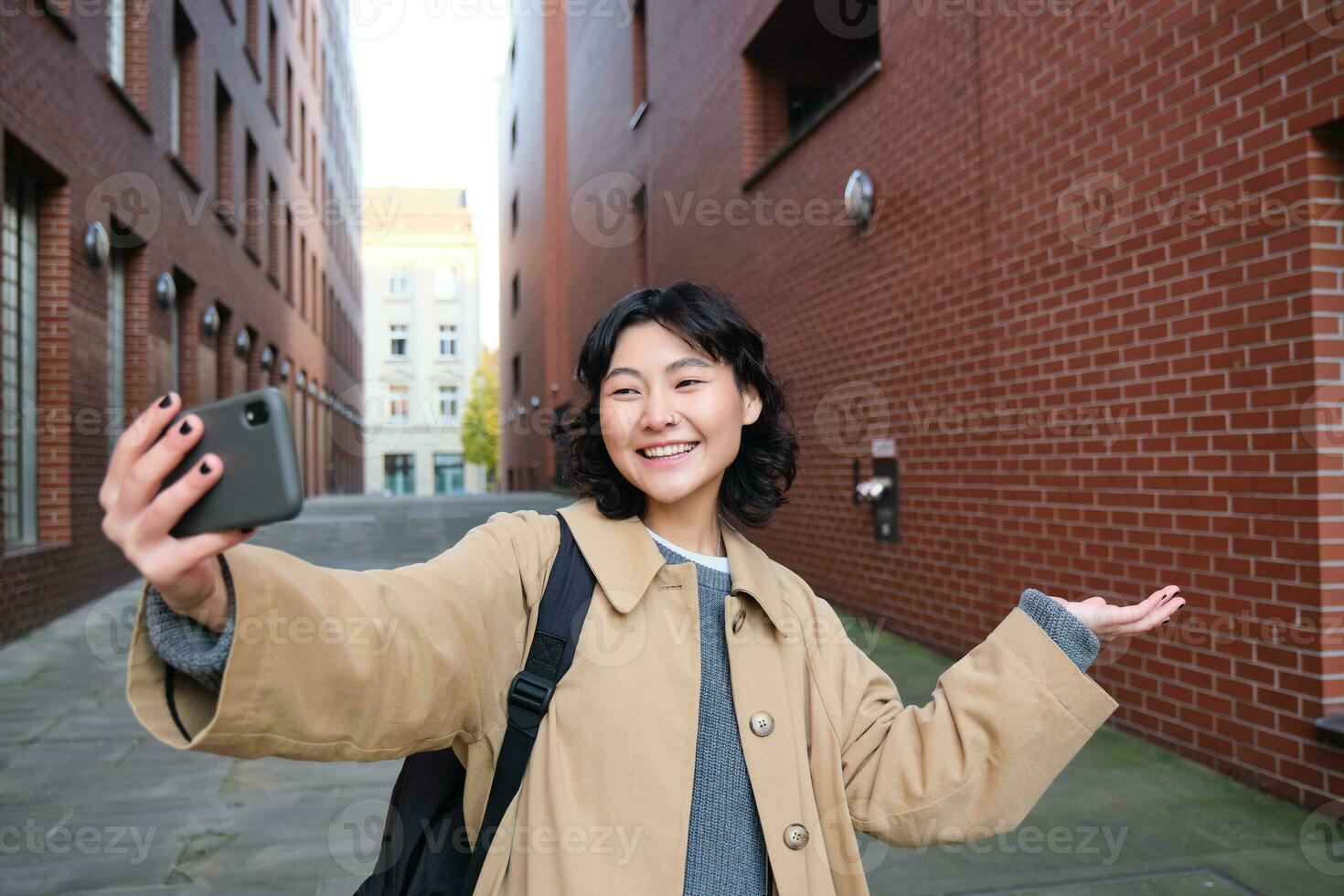 Happy korean girl, tourist takes photos on street, shows smth while records video of herself on smartphone, posing near building and smiling