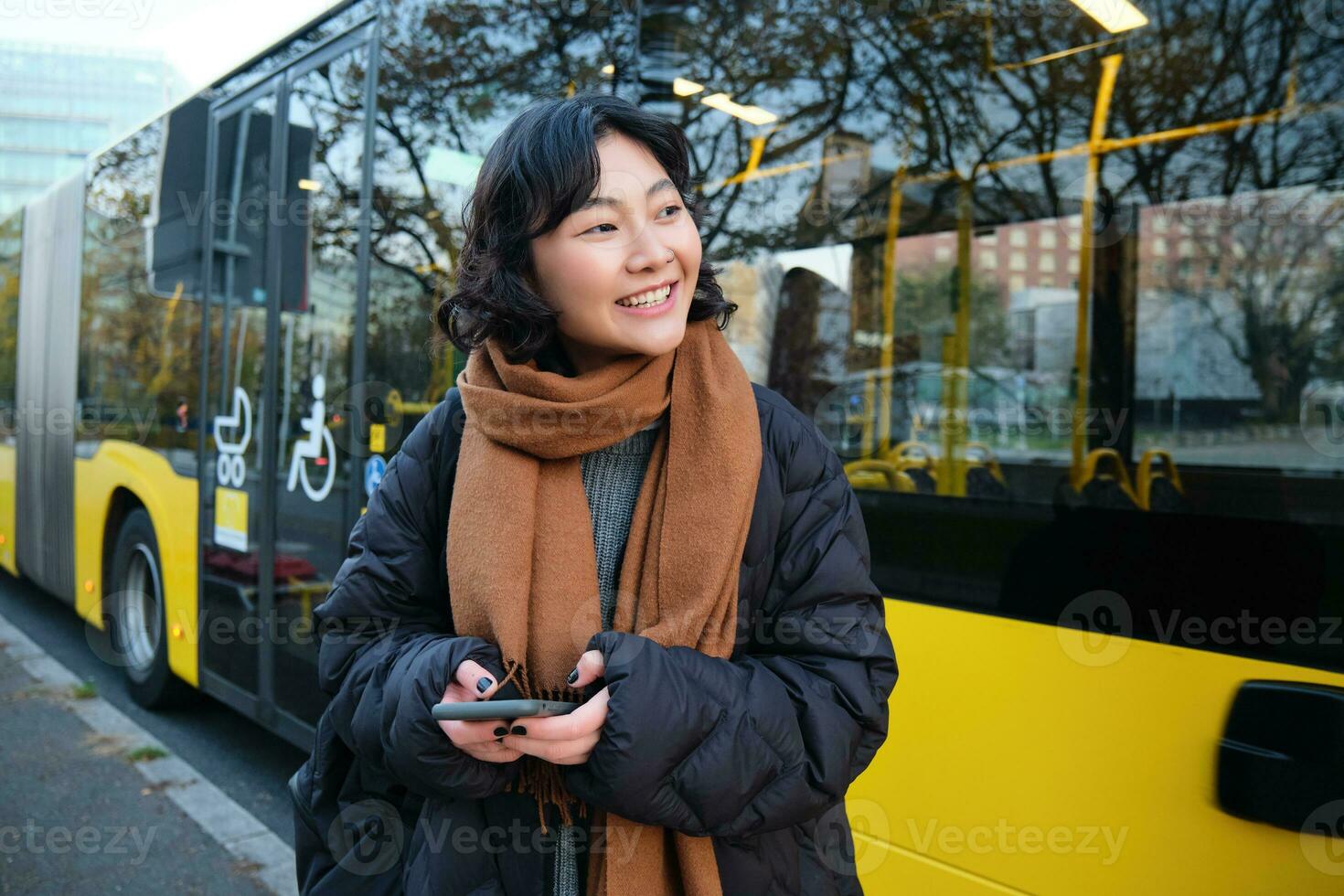 retrato de coreano niña comprando boleto para público transporte en línea, utilizando móvil solicitud en autobús detener, vistiendo invierno ropa foto