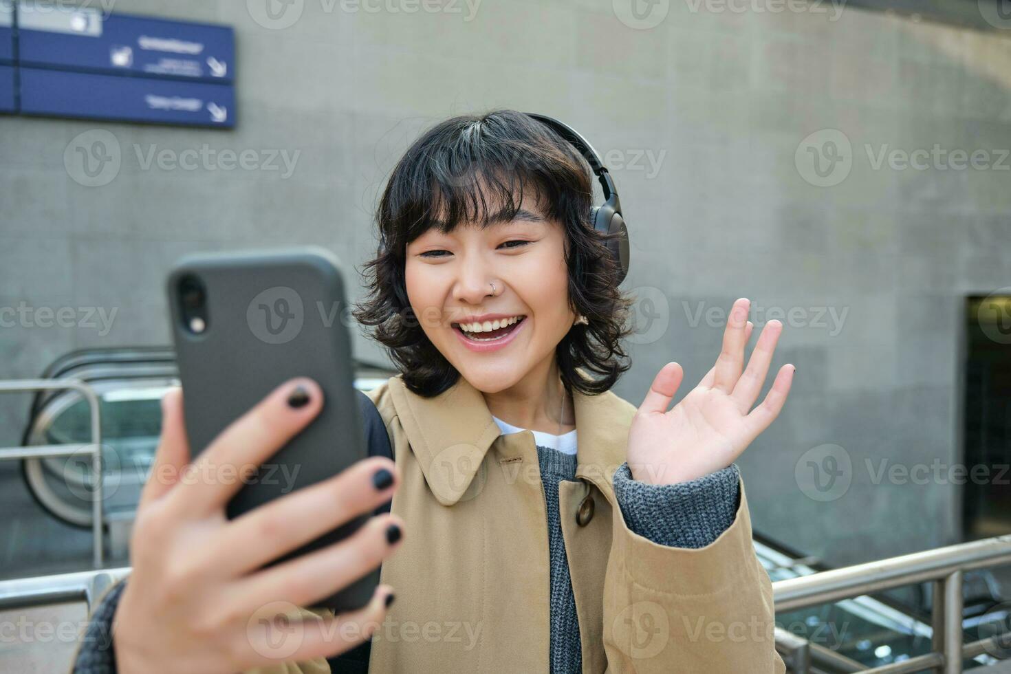 Stylish hipster girl in headphones, says hello to mobile phone camera, waves hand at smartphone, video chats, records a blog, listens music while stands on street photo