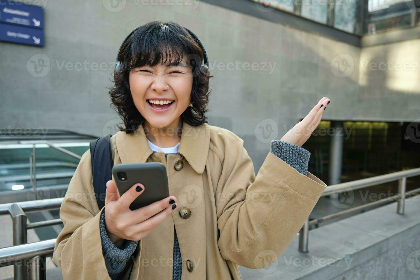Happy asian girl in headphones, looking at mobile phone and celebrating, reading message with good news, screams yes with excitement, stands on street photo