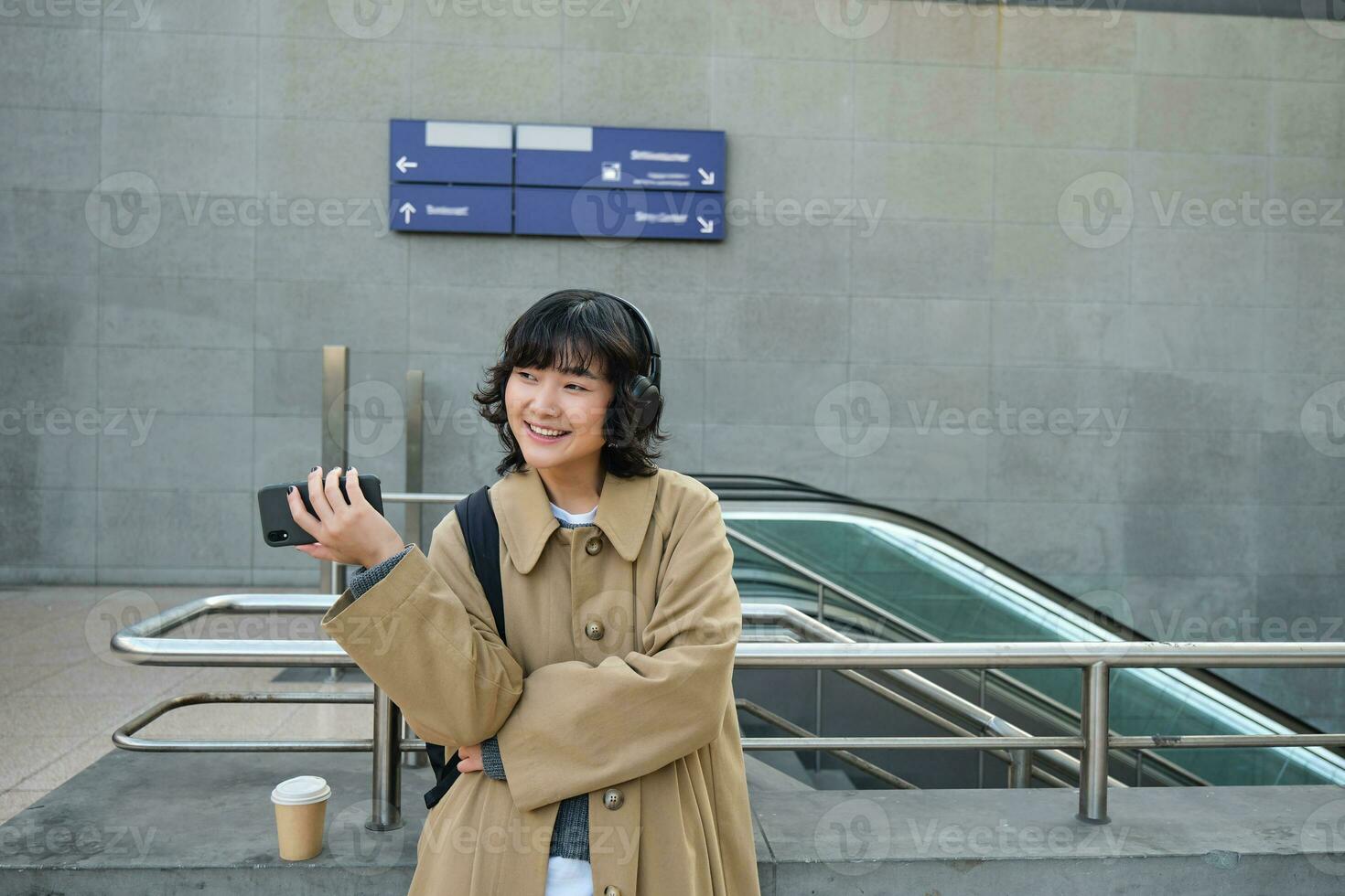 Beautiful brunette girl in trench coat, listens music in headphones and holds mobile phone, waits for someone on street and drinks coffee photo