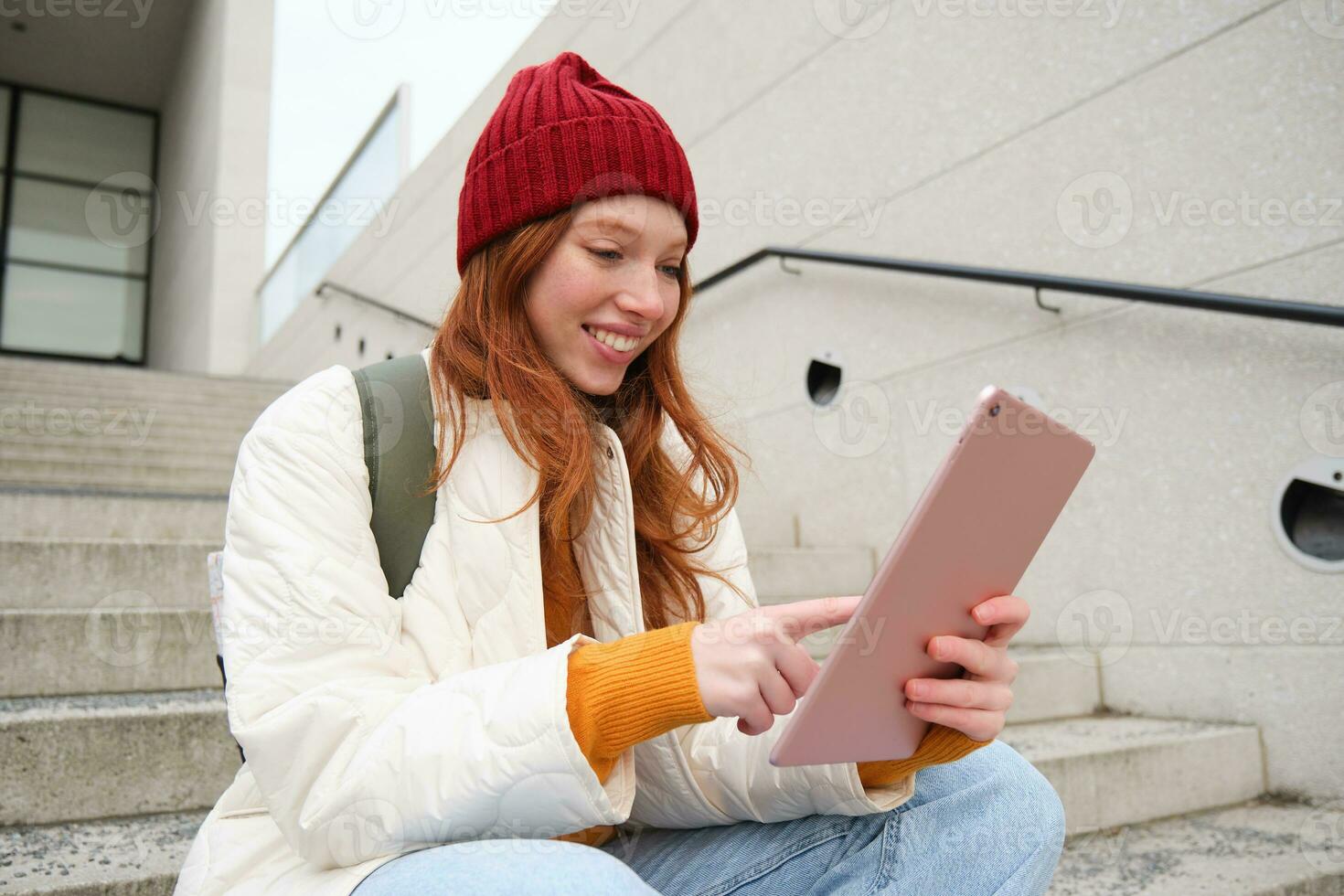 Happy stylish redhead girl, student in red hat, holds digital tablet, uses social media app, searches something online, connects to wifi photo