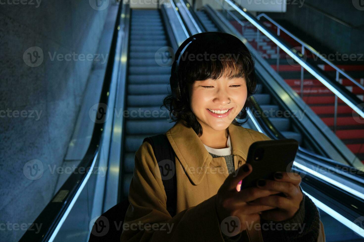 Portrait of girl student in headphones, listens music, commutes, goes down escalator, looks at smartphone with pleased smile, happy face photo