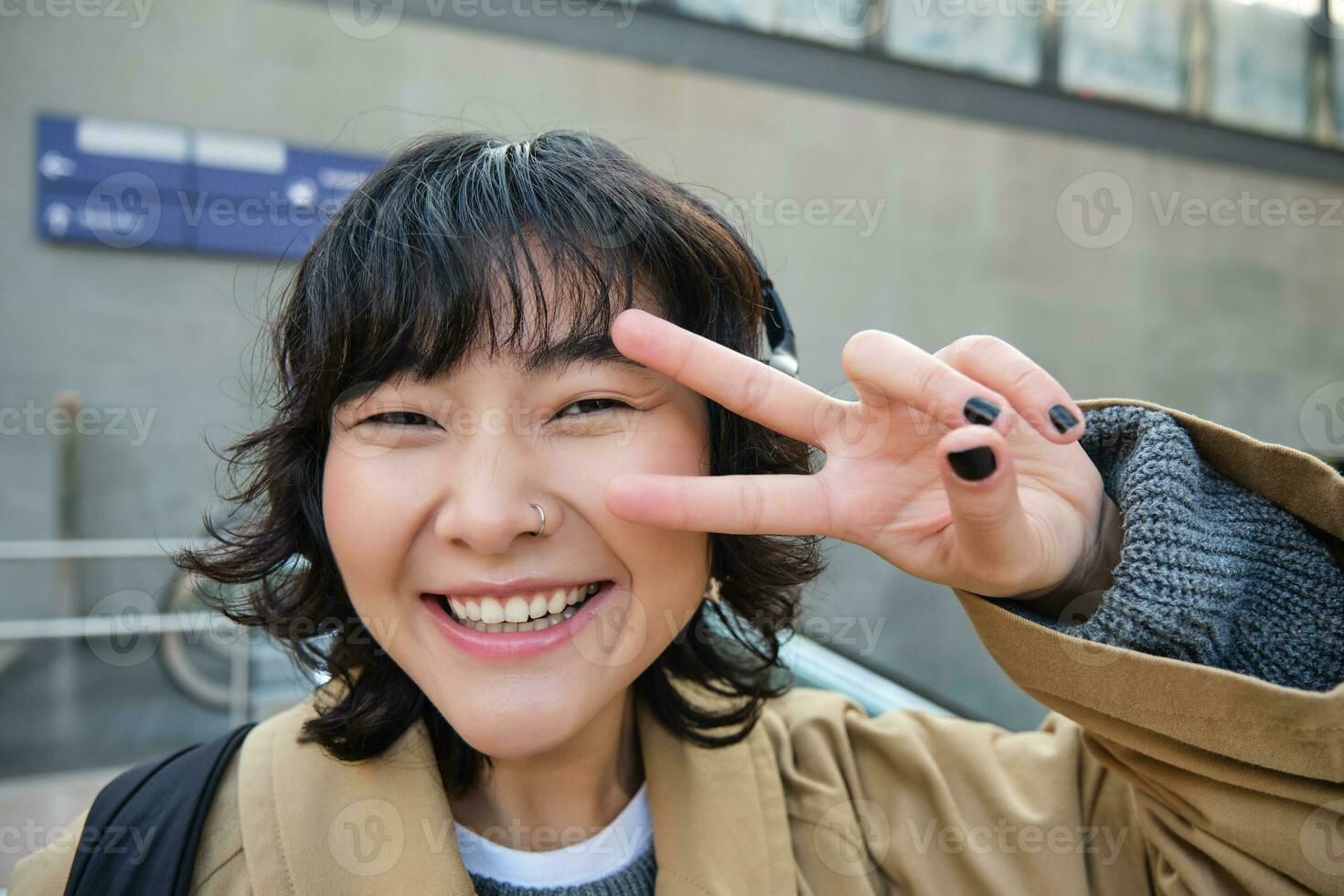 Positive korean girl in headphones, drinks coffee to go, shows peace, v-sign, stands on street and smiles happily photo