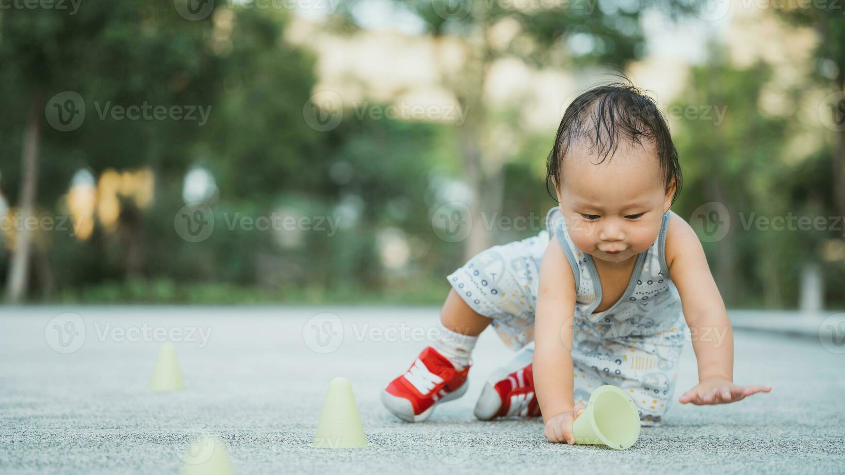 Toddler boy playing with yellow cone marker. Child was about to stand up after falling down. A 1 year old baby is playing at the playground to practice walking. Son is wearing red sneakers. photo