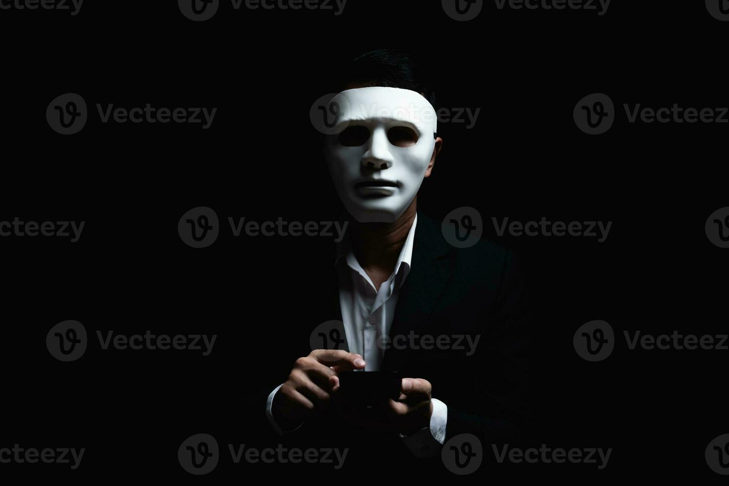 Unknown businessman wearing mask with covered face using mobile phone makes an anonymous call intimidating and threatening the interlocutor on dark background. hacker callcenter concept. photo