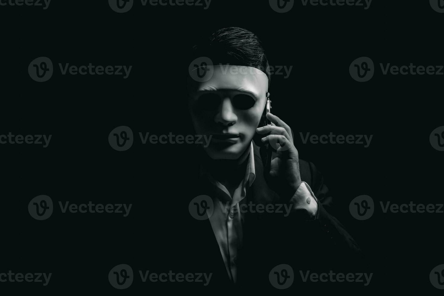 Unknown businessman wearing mask with covered face using mobile phone makes an anonymous call intimidating and threatening the interlocutor on dark background. hacker callcenter concept. photo