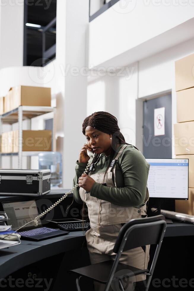 Employee using landline phone to talk with remote client, discussing order detalis before start preparing package for delivery. African american worker wearing industrial overall working in storehouse photo