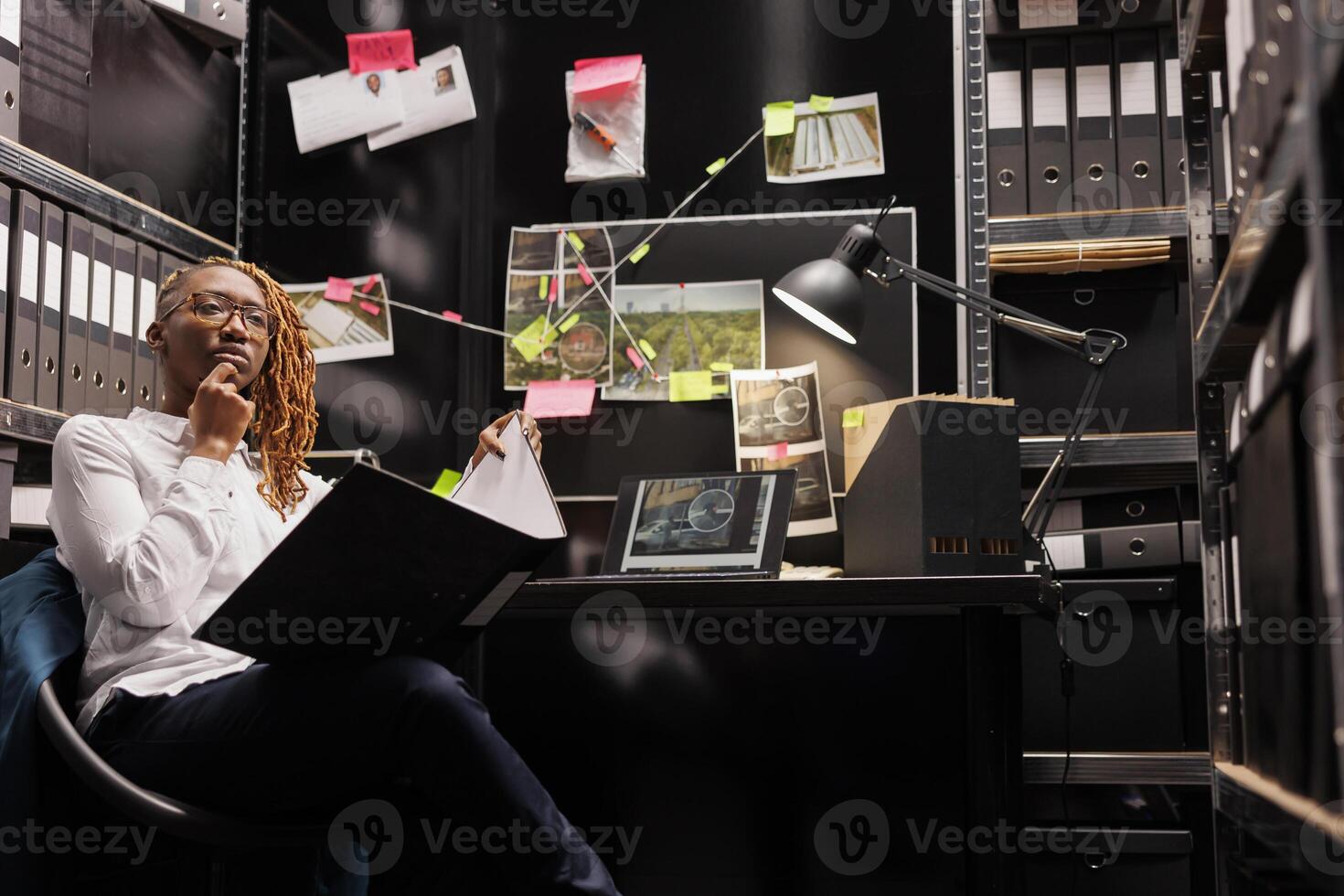 Police detective analyzing criminal case file and rubbing chin while sitting at office table. Pensive woman investigator thinking about crime solving solution while reading csi report photo