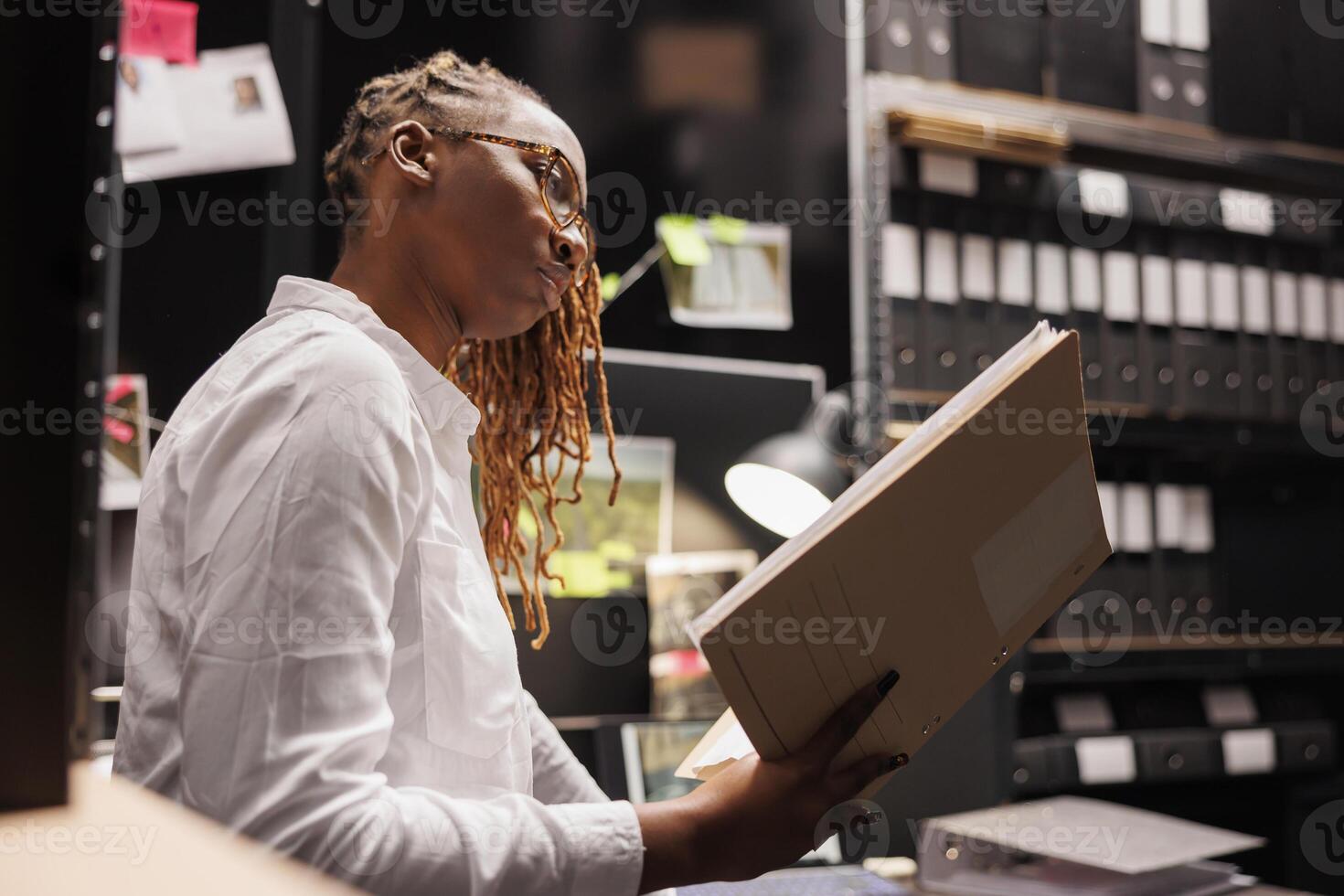 African american woman lawyer analyzing criminal case file in police detective office. Cop studying witness testimony report while sitting at investigator workplace desk at night time photo