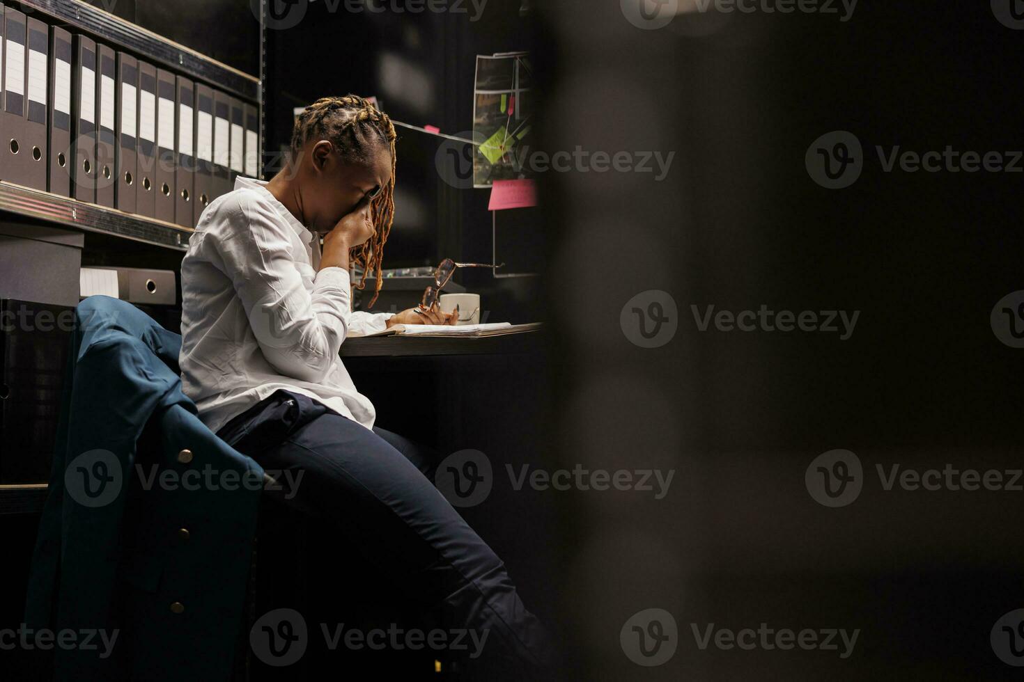 Exhausted police investigator working overtime and rubbing eyes while sitting at workplace desk. Stressed african american woman law enforcement detective in office room at night time photo