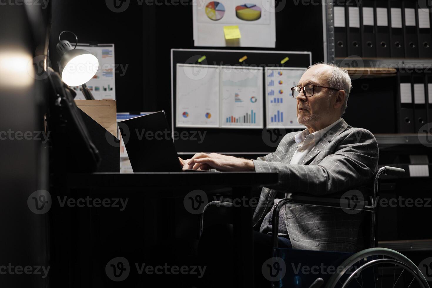 Experienced business manager executive in wheelchair at office desk inspecting quarterly company results analysis data on laptop. Senior with disability on file cabinet office filled with flowcharts photo