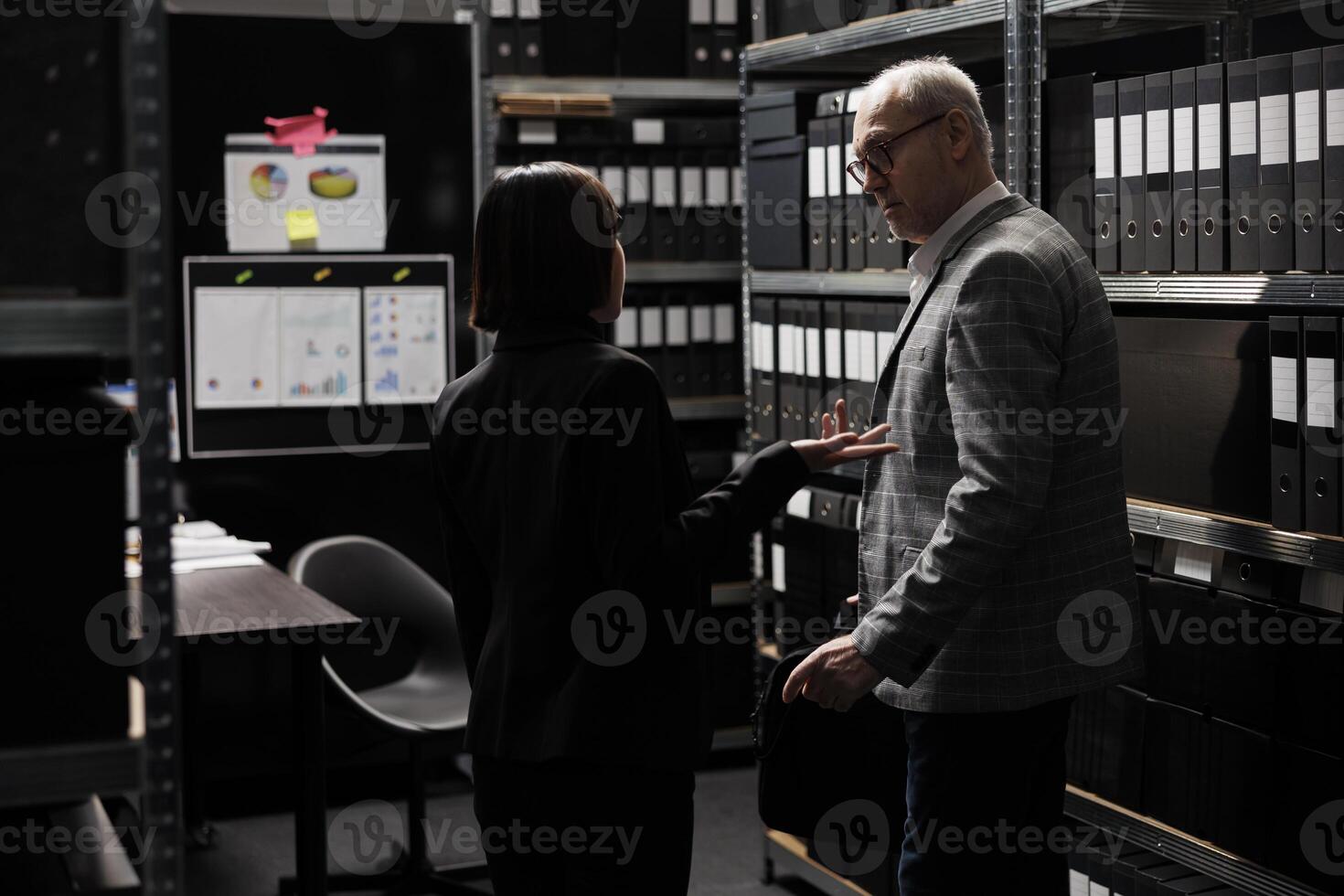 Accountancy executives discussing quarterly profit revenue, analysing performance data. Asian woman and senior businessman in bureaucratic workplace surrounded by documentation folders in cabinets photo